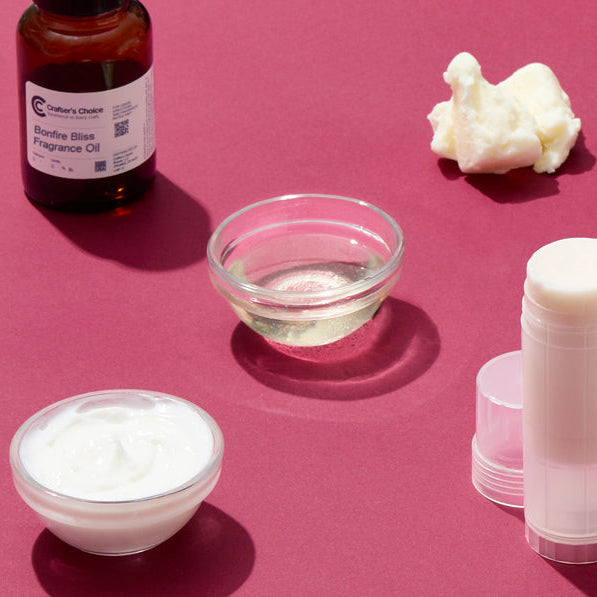 Flat lay of best-selling personal care ingredients and supplies including fragrance oils, wax flakes, lotion, oils, and a clear gel bottle arranged on a bold pink background.