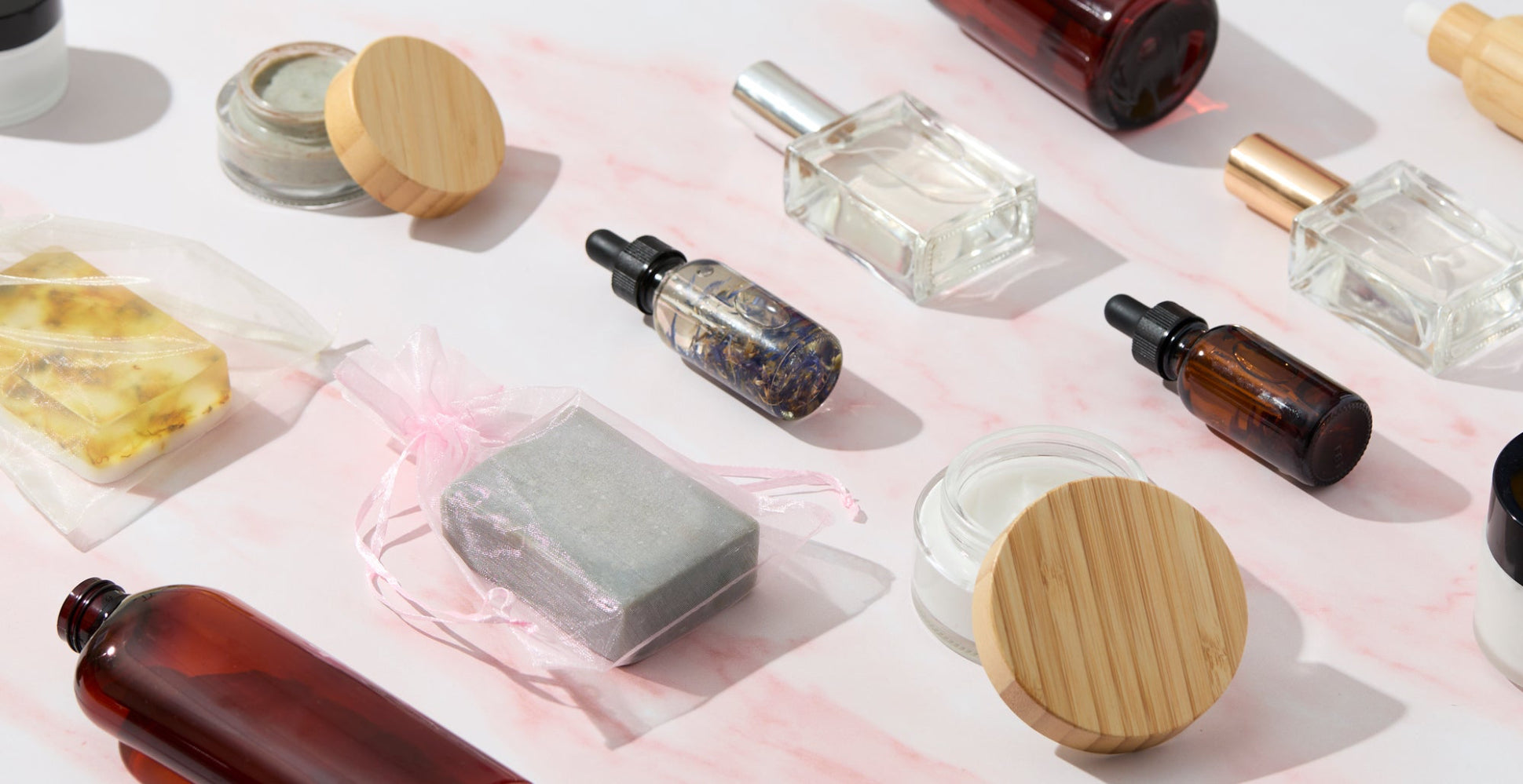 Flat lay of handcrafted soap bars, glass bottles, dropper oils, jars, and packaging supplies arranged on a light pink marble surface, showcasing DIY personal care and cosmetic packaging essentials.