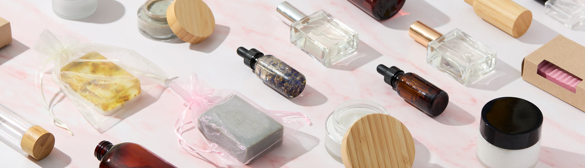 Flat lay of handcrafted soap bars, glass bottles, dropper oils, jars, and packaging supplies arranged on a light pink marble surface, showcasing DIY personal care and cosmetic packaging essentials.