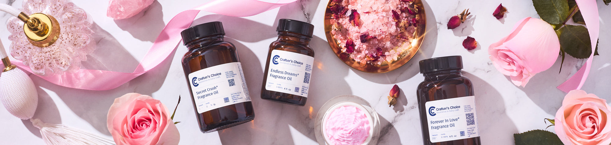 Flat lay of amber glass fragrance oil bottles labeled “Secret Crush,” “Endless Dreams,” and “Forever In Love,” surrounded by pink roses, ribbon, bath salts, and a candle on a marble surface.