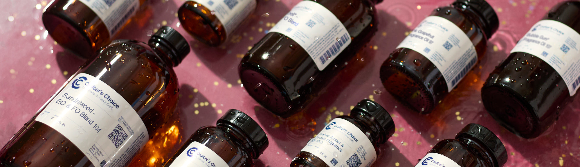 Assorted Crafter’s Choice fragrance oil bottles scattered on a pink surface with water droplets and reflective highlights.