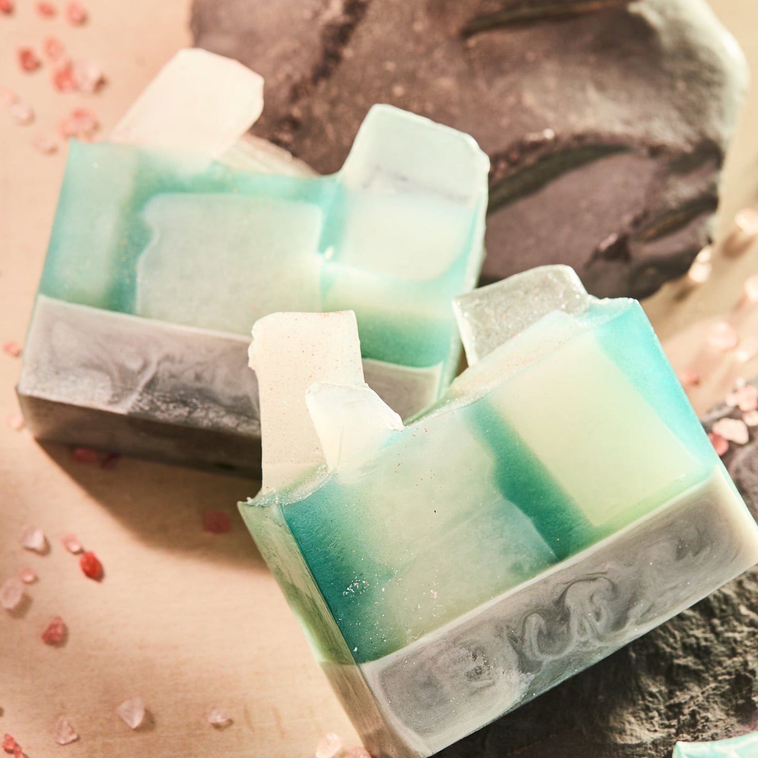 Handcrafted mineral-style soap bars with layered shades of seafoam green, white, and charcoal arranged on textured stone. Scattered mineral salts and warm lighting highlight the soap’s translucent layers and natural, artisanal finish.