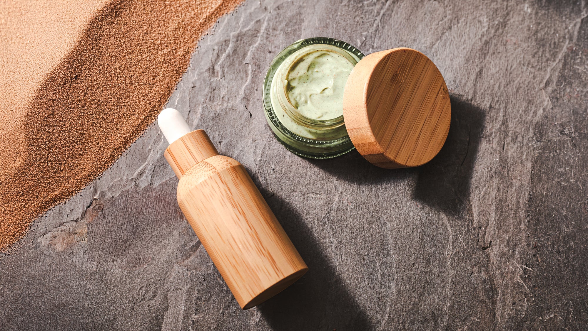 Bamboo skincare packaging arranged on a textured stone surface, featuring a wooden dropper bottle and an open glass jar of green cream with a bamboo lid. Warm lighting and scattered clay powder create a natural, spa-inspired scene.