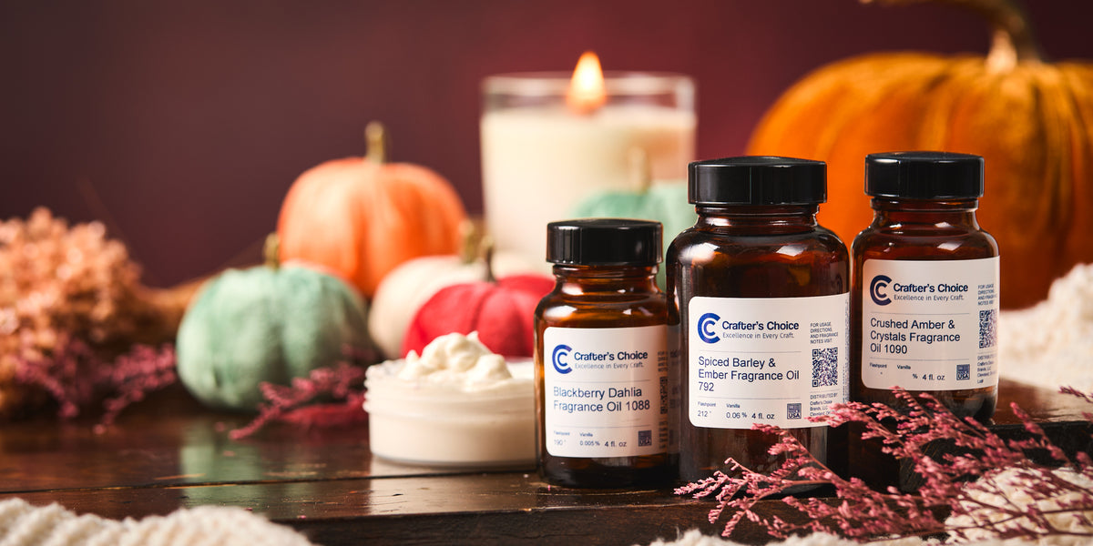 Autumn-themed display featuring four amber bottles of Crafter's Choice fragrance oils, a jar of white whipped body butter, colorful mini pumpkins, a lit candle, dried florals, and a large pumpkin on a rustic wooden surface.