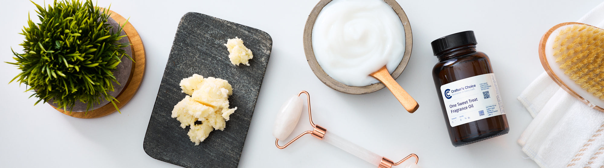 Flat lay of personal and home care making supplies including lotion base, a jade roller, a fragrance oil bottle, and a dry brush on a white background.