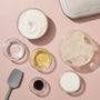 Top-down view of soap-making ingredients arranged on a soft pink background, including a bowl of white whipped base, clear liquid in a glass bowl, golden oil, translucent soap cubes, white granules, dark red liquid colorant, a gray spatula, and a metal tin lid.
