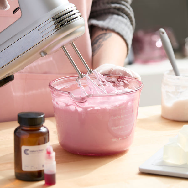 Person wearing gloves and a pink apron uses a handheld mixer to whip a light pink soap mixture in a clear glass measuring bowl on a wooden table. Nearby are a small amber bottle, red liquid colorant, soap cubes on a digital scale, and a spatula in a jar.