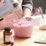 Person wearing gloves and a pink apron uses a handheld mixer to whip a light pink soap mixture in a clear glass measuring bowl on a wooden table. Nearby are a small amber bottle, red liquid colorant, soap cubes on a digital scale, and a spatula in a jar.