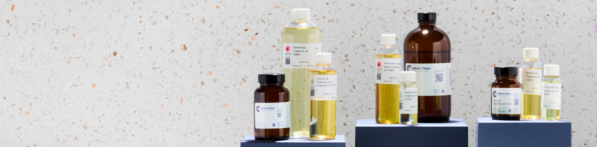 Assorted fragrance oil bottles in various sizes displayed on blue pedestals against a light terrazzo background, featuring clear and amber containers filled with golden and pale yellow oils.