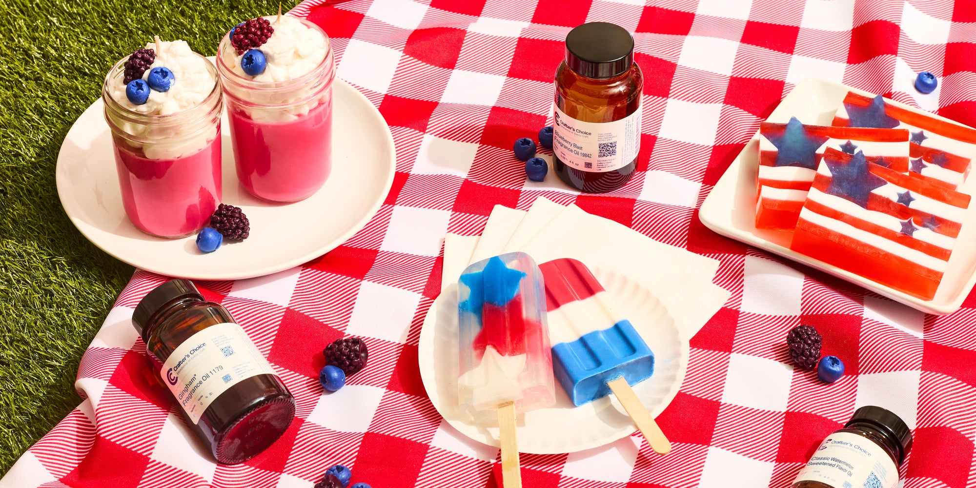 Red, white, and blue patriotic picnic setup with layered star-and-stripe gelatin treats, berry-topped pink parfaits, popsicles, and decorative stars on a red checkered blanket outdoors.