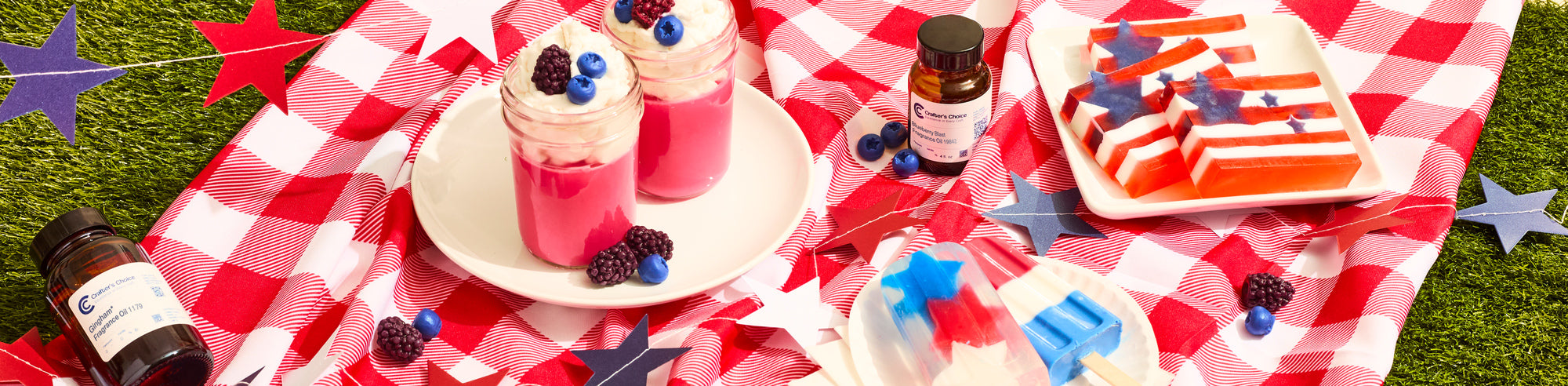 Red, white, and blue patriotic picnic setup with layered star-and-stripe gelatin treats, berry-topped pink parfaits, popsicles, and decorative stars on a red checkered blanket outdoors.