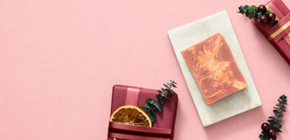 
Cranberry orange soap with peach and cream swirls on a marble tray, surrounded by wrapped gifts, dried orange, and greenery on a pink background.