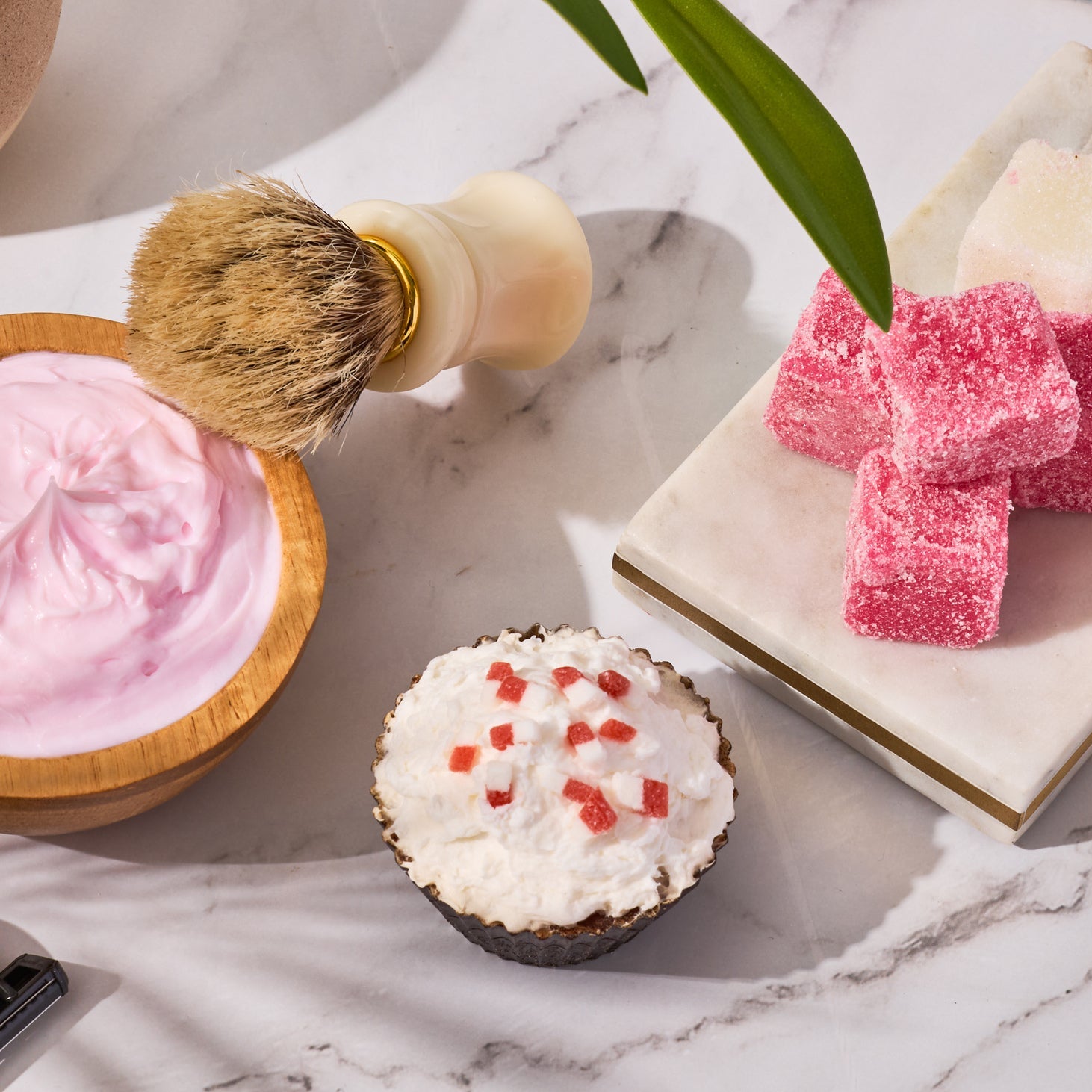 Flat lay of a spa-style shaving and bath scene with a bowl of pink whipped cream, shaving brush, sugar scrub cubes, a creamy soap cupcake, and greenery on a marble surface.