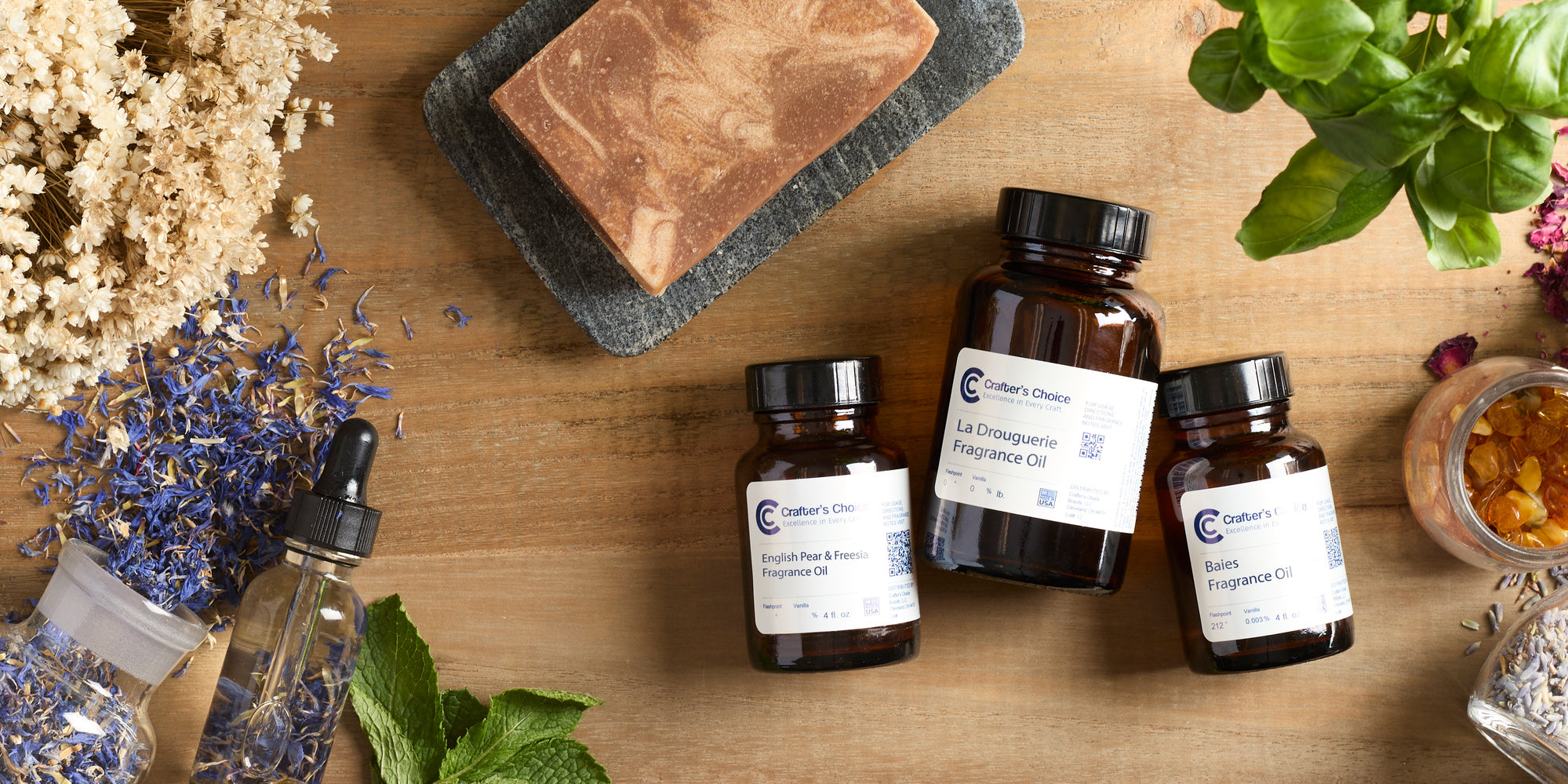 Close-up flat lay of soap-making setup: marbled bar of soap on a stone dish, amber fragrance oil bottles with labels, dropper bottle, dried flowers, lavender, and fresh basil and mint on a wooden surface.