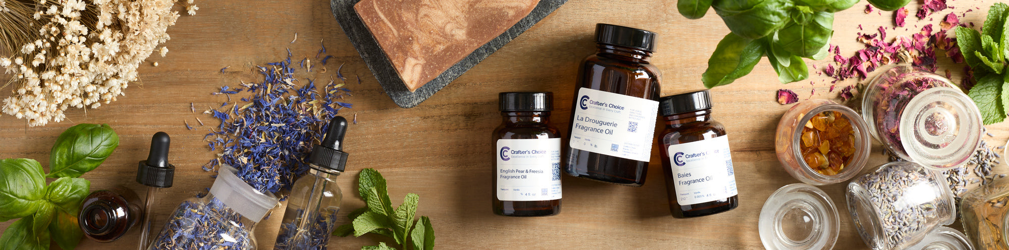 Flat lay of soap-making ingredients on wooden table: amber bottles of fragrance oil, dropper bottles, glass jars, dried lavender and flowers, basil and mint leaves, and a marbled bar of soap.