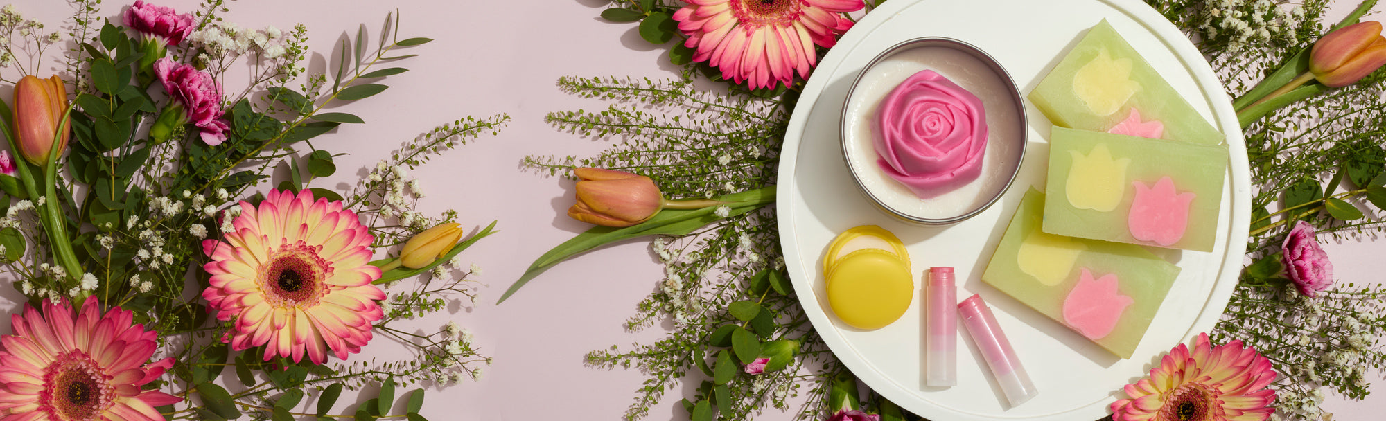 Flat lay of spring flowers surrounding a white tray with handmade soaps, a rose-shaped candle, yellow balm, and pink lip balms on a soft pink background.