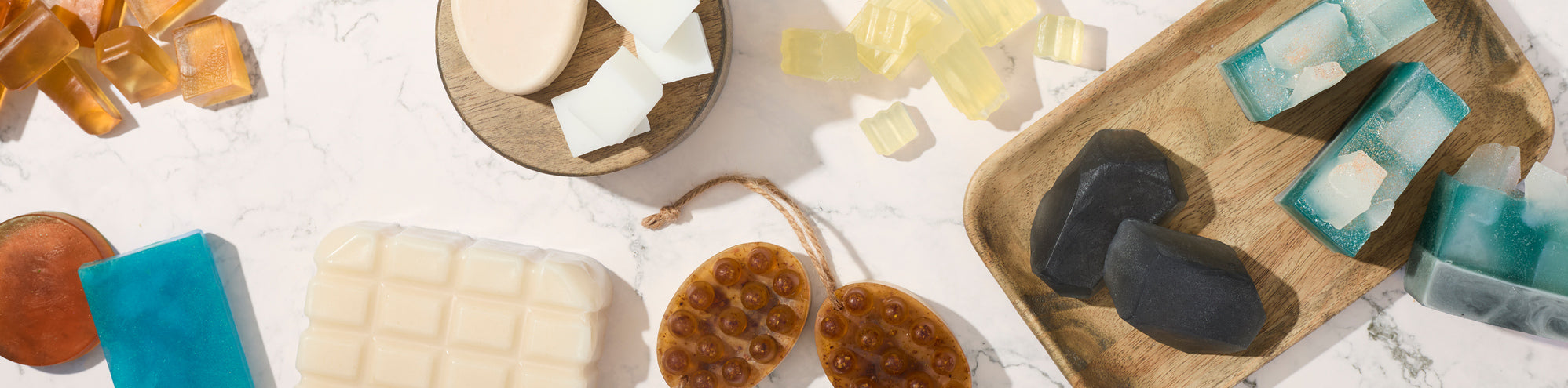 Assorted handmade soaps arranged on marble and wooden trays, including blue and teal bars, translucent amber cubes, a white block soap, black geometric soaps, and a honeycomb-textured exfoliating soap with a rope loop.