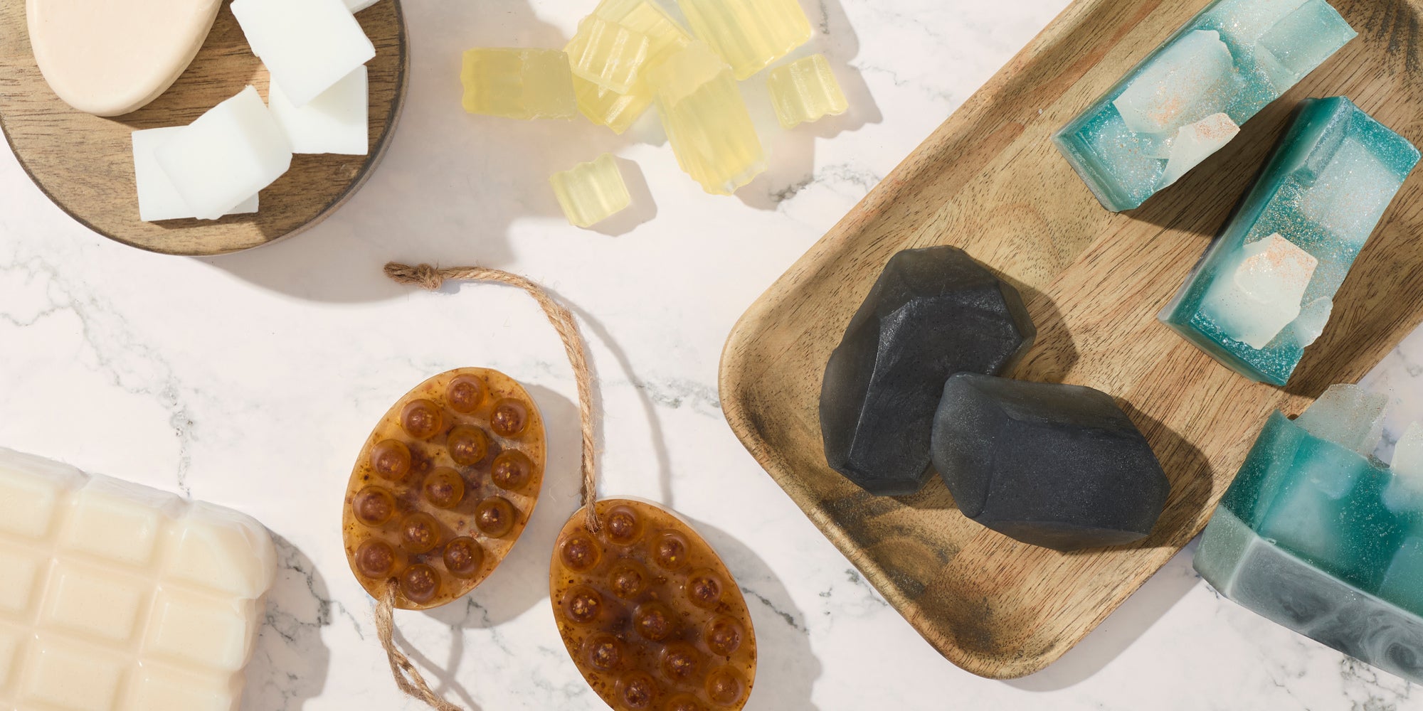Assorted handmade soaps arranged on marble and wooden trays, including blue and teal bars, translucent amber cubes, a white block soap, black geometric soaps, and a honeycomb-textured exfoliating soap with a rope loop.