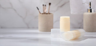 Two translucent deodorant tubes filled with cream-colored balm displayed on a bathroom countertop with blurred toiletries in the background.