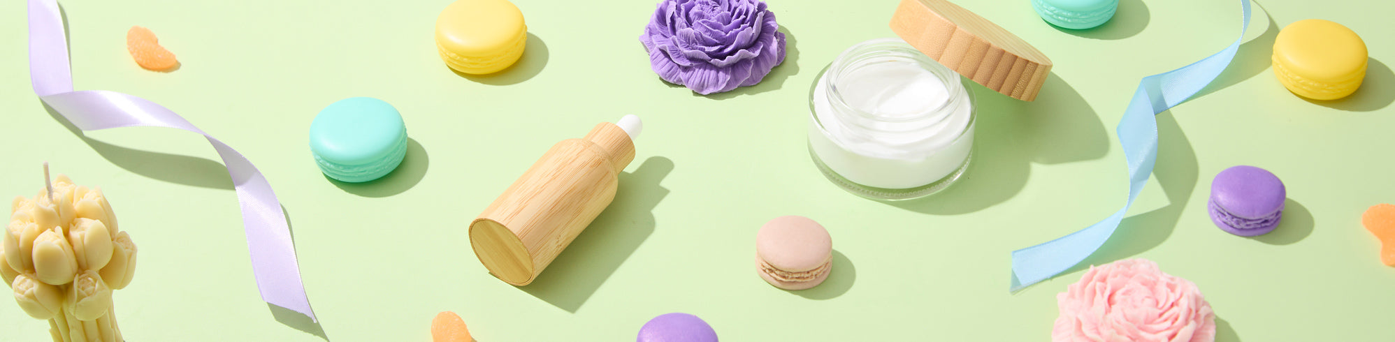 Flat lay of pastel items on light green background: colorful macarons, ribbons, floral-shaped soaps or candles, a glass jar of cream, and a wooden dropper bottle arranged with soft shadows.