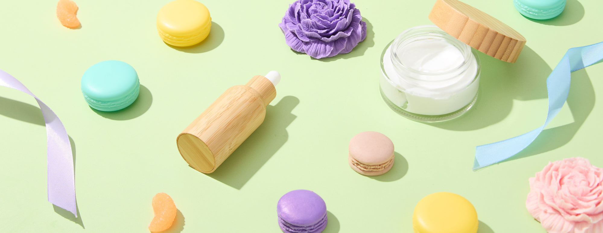 Flat lay of pastel items on light green background: colorful macarons, ribbons, floral-shaped soaps or candles, a glass jar of cream, and a wooden dropper bottle arranged with soft shadows.