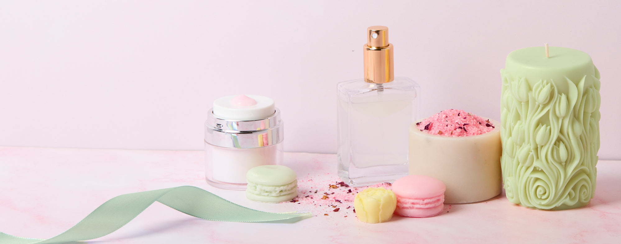 Pastel-themed arrangement of beauty and decor items: clear perfume bottle with gold cap, cream jar, green carved candle, pink bath salt candle, and pastel macarons on a soft pink surface with scattered petals