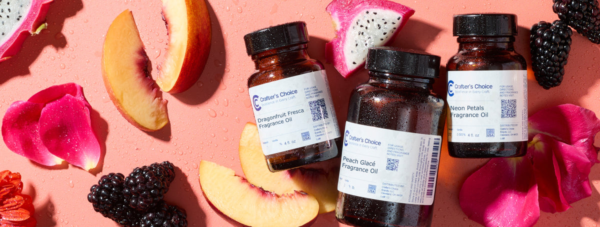 Three amber glass fragrance oil bottles labeled Crafters Choice—Dragonfruit Fresca, Peach Glacé, and Neon Petals—surrounded by sliced peach, dragonfruit, blackberries, pink petals, and flowers on a coral background with water droplets.