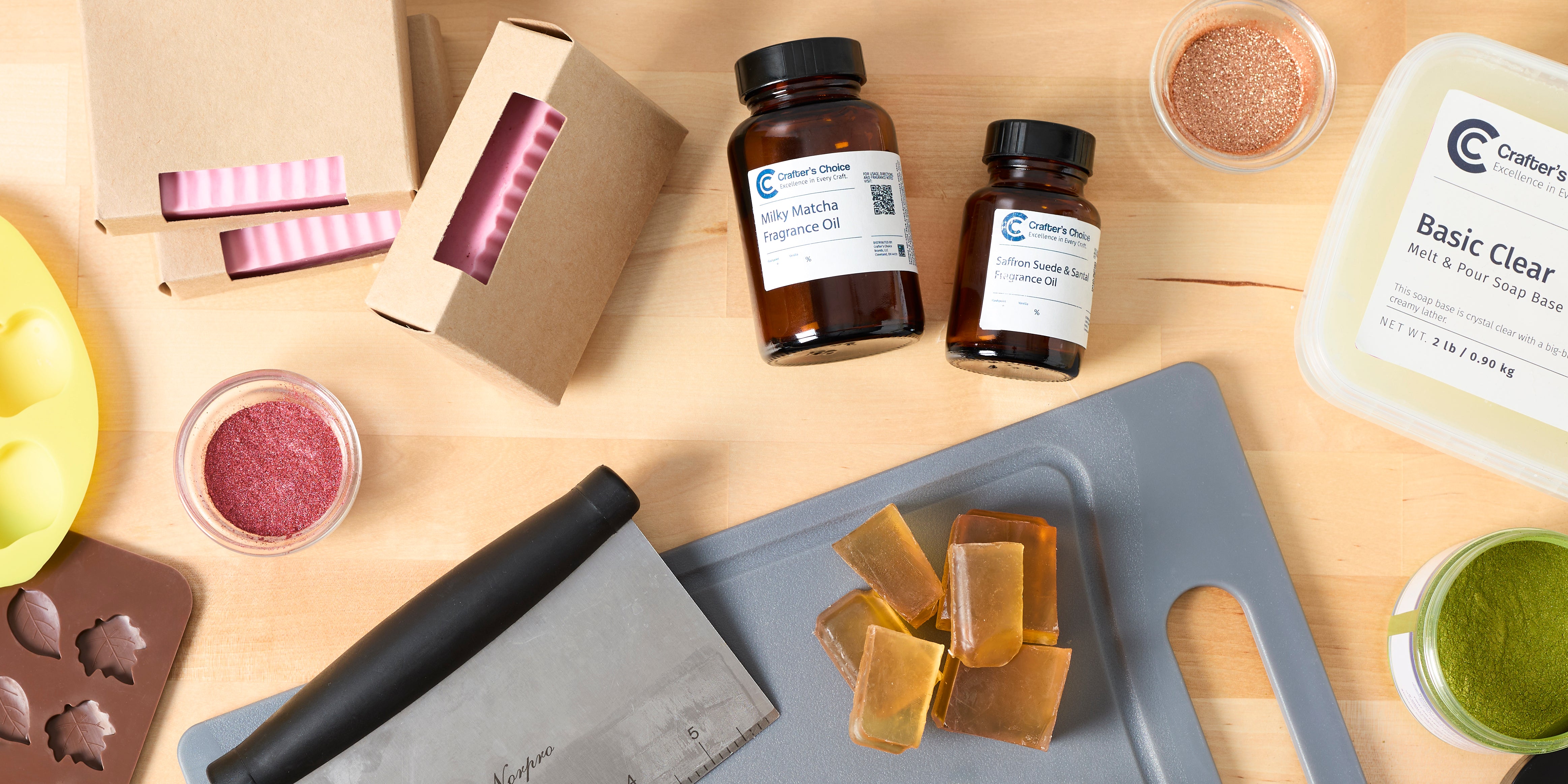 Flat lay of soap making supplies including silicone leaf and apple molds, handcrafted soap bars in kraft boxes, fragrance oils, clear melt-and-pour soap base, mica powders, soap cutter, and soap cubes on a wooden surface.