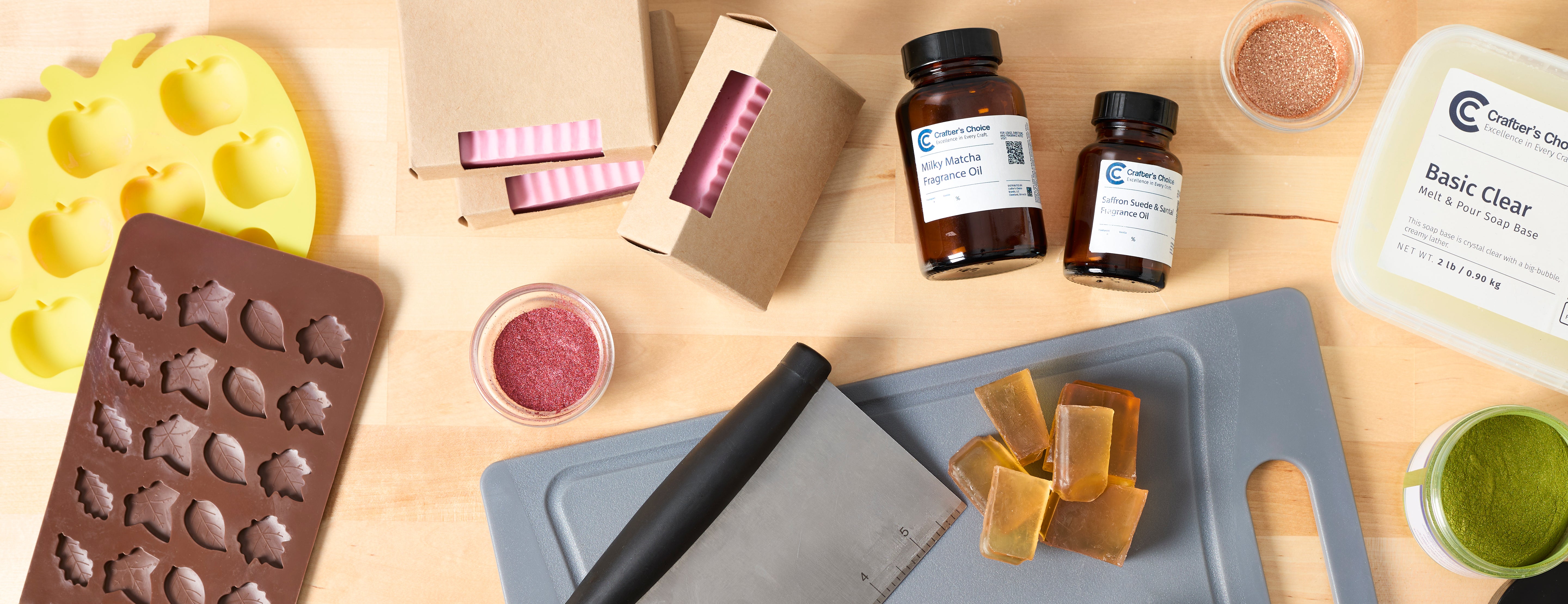Flat lay of soap making supplies including silicone leaf and apple molds, handcrafted soap bars in kraft boxes, fragrance oils, clear melt-and-pour soap base, mica powders, soap cutter, and soap cubes on a wooden surface.