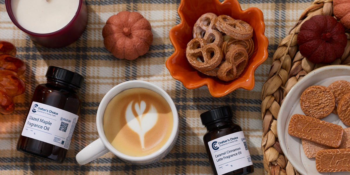 Autumn flat lay with fragrance oil bottles, a latte with leaf art, pumpkin-shaped décor, candles, and assorted cookies arranged on a plaid blanket and woven placemat.