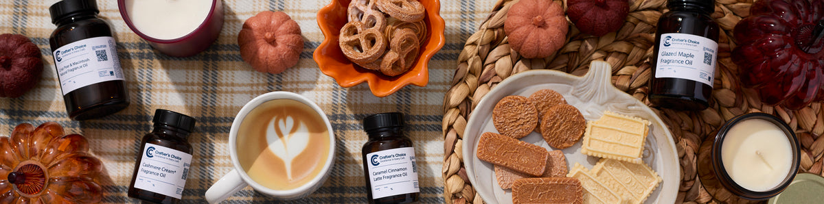 Autumn flat lay with fragrance oil bottles, a latte with leaf art, pumpkin-shaped décor, candles, and assorted cookies arranged on a plaid blanket and woven placemat.
