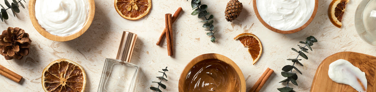 Skincare products and perfume arranged with dried orange slices, cinnamon sticks, pinecones, and eucalyptus on a cream surface, creating a warm, natural seasonal display.