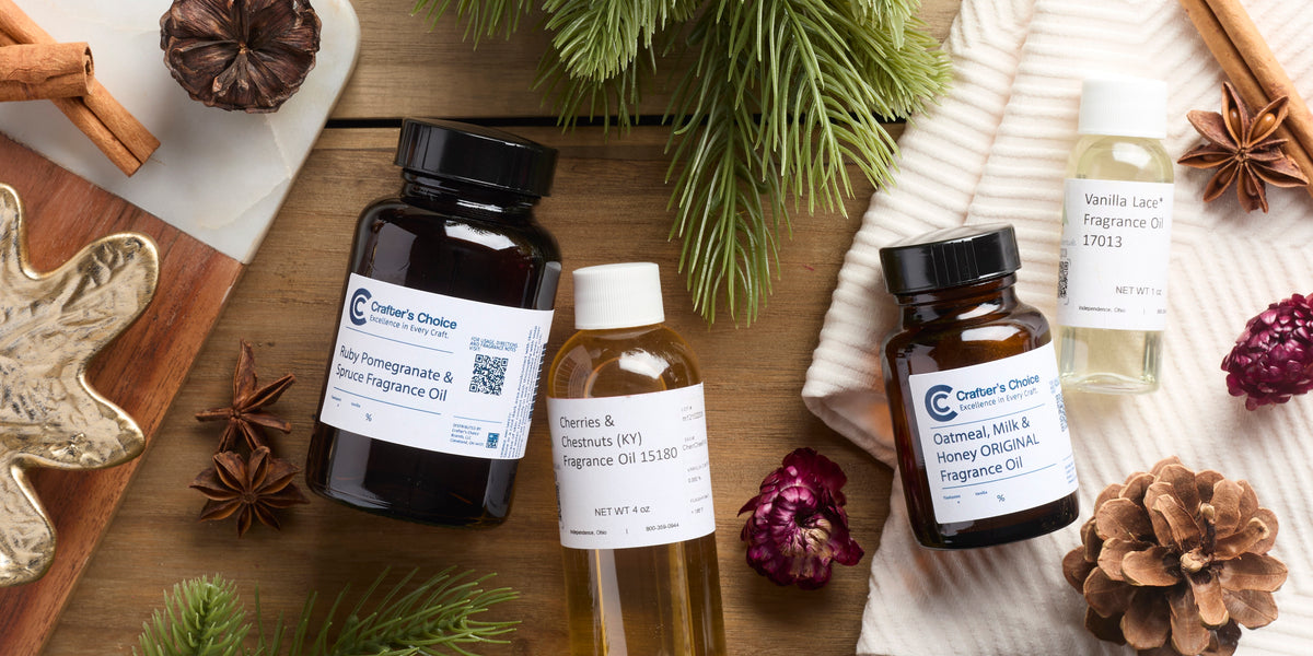 Assorted fragrance oil bottles arranged on a rustic wooden surface with pine branches, pinecones, cinnamon sticks, star anise, dried flowers, a white knit blanket, and a gold leaf-shaped dish.