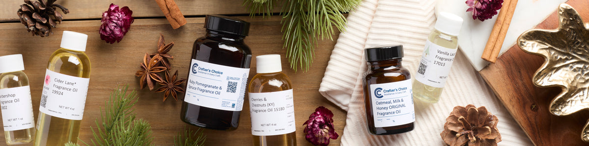 Assorted fragrance oil bottles arranged on a rustic wooden surface with pine branches, pinecones, cinnamon sticks, star anise, dried flowers, a white knit blanket, and a gold leaf-shaped dish.