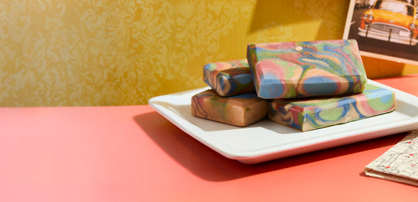 Four colorful handcrafted soap bars with marbled blue, pink, green, and tan swirls stacked on a white square plate against a vibrant yellow patterned backdrop and coral pink surface, with a vintage car photo in the background.